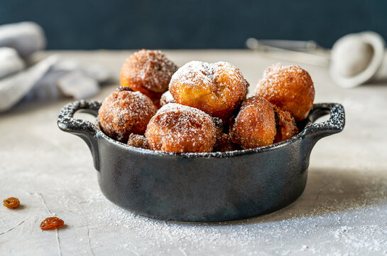 Carnival fritters or balls are Italian sweets dropped with sugar powder in a black pot on concrete table with dark background, strainer, strainer, raisins