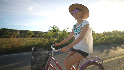 Closeup side view of pretty mature senior woman biking away from the sun, backlit, wearing sunglasses, hat and ethnic clothes in a tropical setting.