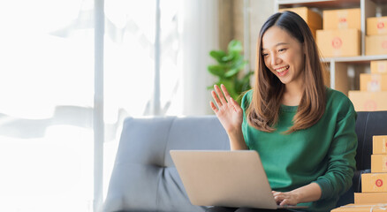 Fototapeta premium Start a small business, young asian woman live online shopping with packing boxes and checking online purchases on digital laptop for products to be delivered to customers.