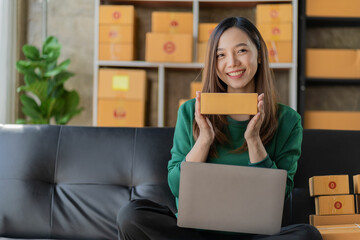 Concept of small business owner, attractive young asian businesswoman holding yellow box working happily with laptop in online shopping at home preparing parcel delivery in sme supply chain