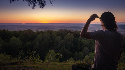 A young man watching over forest landscape from a view point and touches his base-cap. The sky over the horizon is orange after sunset. - obrazy, fototapety, plakaty