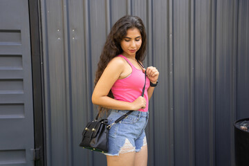 Young attractive fashionable indian woman wearing casual jeans short outfit and handbag standing outdoor.