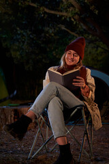 Serene caucasian woman at camping sitting outside during the night reading a book, disconnecting and enjoying the nature. Vertical photo with Copy space.