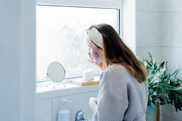 Young woman in bathrobe looking in the mirror and removing natural clay mask with loofah pad from...