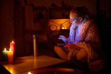Young girl in warm plaid sitting at home and working on laptop in the evening with candle light. No electricity, blackout. Adjusting to unstable electricity service. Lifestyle in Ukraine