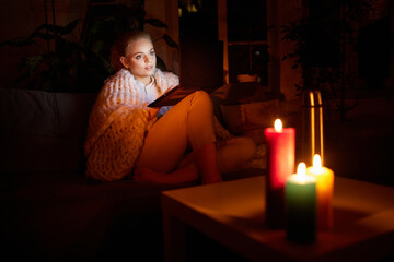 Adjusting to unstable electricity. Young girl, freelancer sitting on sofa at home in evening and working, watching movie on laptop with candle light. Blackout. Concept of power outage