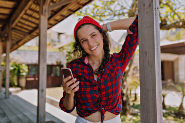 Adorable happy stylish European girl in knitted cap and striped shirtpisng to camera with happy smile against wooden wall an during smartphone 