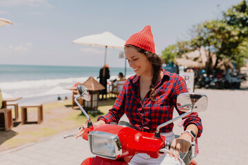 Smiling happy girl in cap and striped shirt sitting on red bike with broadly smile against blue sky...