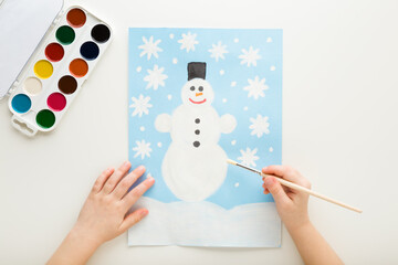 Little child hand holding paint brush and drawing snowman and snowflakes on light blue paper on...