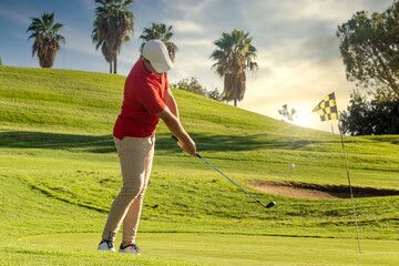 golfer shooting an amazing golf ball from the fairway to the flagstick on the green. Side view of golfer hitting a golf ball with approach shot. Golf course at sunset copy space