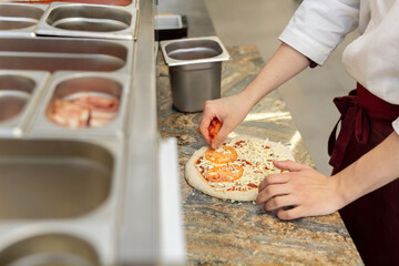 The chef in the restaurant kitchen adds a fresh tomato to the pizza.