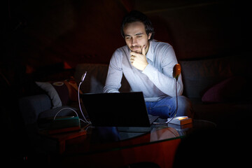 Adjusting to unstable electricity. Man, freelancer sitting on sofa at home in evening and working...