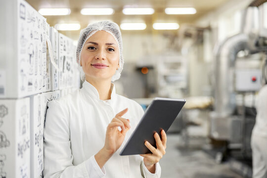 A satisfied food factory supervisor is monitoring works on tablet while smiling at the camera.