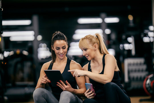 A happy sportswoman is pointing at the tablet while her female personal trainer is showing her progress in a gym.