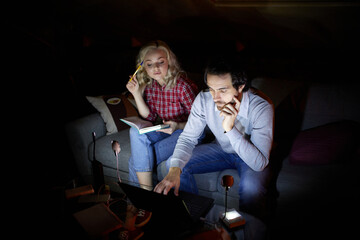 Man and woman, couple sitting on sofa at home in the evening and studying, working with little...