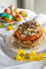 Traditional Russian Easter cake - kulich with royal icing and chocolate flakes on white tablecloth background with colored eggs. Yellow forsythia is on the front view.