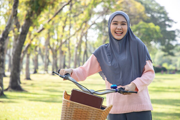 Asian muslim woman enjoying weekend with favorite book at park, lead the bike and looking at camera. Education and relax after riding bicycle. Bright tone and sunny.