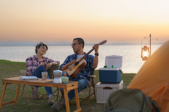 Asian couple singing and playing guitar in their camping area with lake in the background during sunset.