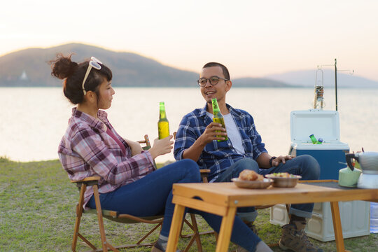 Asian couple drinking beer from bottle in their camping area with lake in the background during sunset.