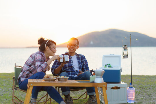 Asian couple having a cup of coffee or water at their camping site with lake in the background during sunset