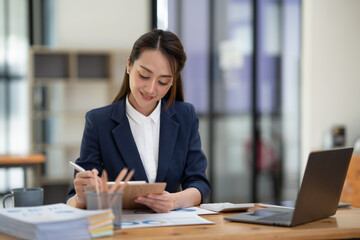 Beautiful attractive Asian businesswoman holding a clipboard of documents sitting analytically analyzing various marketing management information the finances were at her desk in the office at ease. © crizzystudio