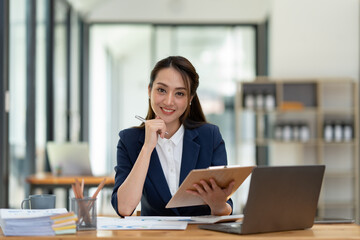 Beautiful attractive Asian businesswoman holding a clipboard of documents sitting analytically analyzing various marketing management information the finances were at her desk in the office at ease. © crizzystudio