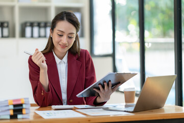 Beautiful attractive Asian businesswoman holding a clipboard of documents sitting analytically analyzing various marketing management information the finances were at her desk in the office at ease. © crizzystudio