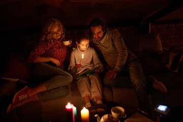 Cozy family evening. Mother, father and little girl sitting on sofa without electricity and reading...