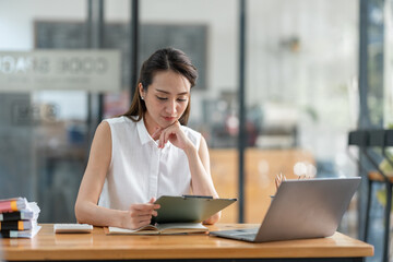 Beautiful attractive Asian businesswoman holding a clipboard of documents sitting analytically analyzing various marketing management information the finances were at her desk in the office at ease. © crizzystudio