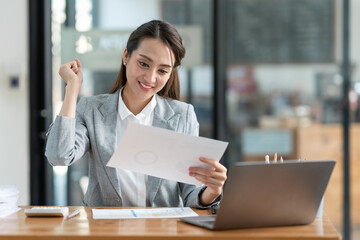 Asian businesswoman sitting happily in office holding detailed report paper analyzing data accuracy in financial matters management concept.