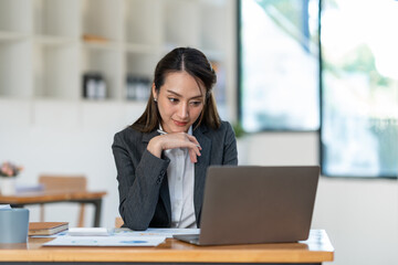 Young Asian businesswoman holding a pen sitting and analyzing the details of management information Finance is happy at the desk in the office. © crizzystudio