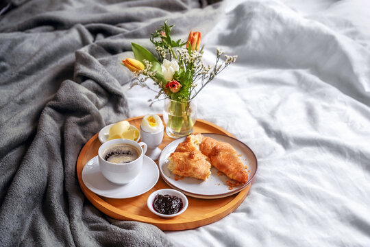 Wooden breakfast tray with croissant, coffee, jam, egg and a fresh flower bouquet served on bed with light linen and gray blanket, holiday or weekend morning, copy space