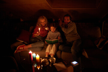 Cozy family evening. Mother, father and little girl sitting on sofa without electricity and reading...