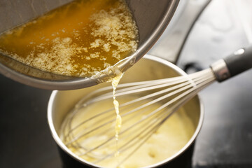 Cooking process of hollandaise sauce, pouring melted butter into the pot with egg mixture, whisking all the time at low temperature to get a creamy texture, selected focus - 567390474