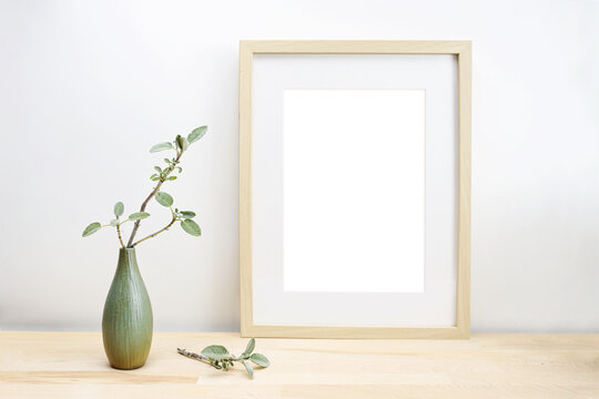 Empty wooden picture frame with passe-partout as mock-up and a vase with sage twig on a light table or desk in front of a white wall, minimalist interior decoration, copy space