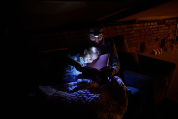 Father with his little daughter sitting on sofa at home in the evening and reading book with...