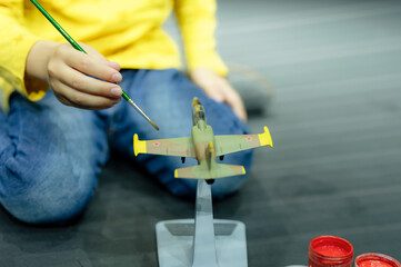 candid little six year old kid boy painting or colorize or colour model airplane with gouache paints while squatting on floor studio illuminated by white yellow bright light
