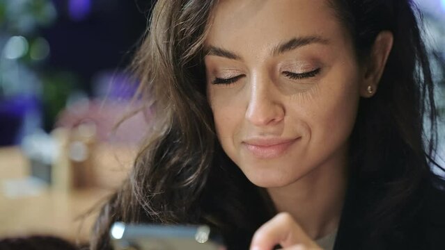 Caucasian female with long brown hair sitting inside restaurant, sipping coffee, smiling, and typing on her smartphone. Close-up, slow motion.