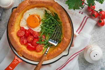 Dutch baby pancake. Fresh homemade Dutch Baby pancake with fried egg, tomato and green arugula in reed cast-iron pan on light gray concrete rustic table. Flat lay, close up.