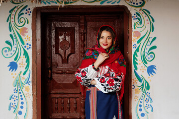 Fototapeta na wymiar Charming smiling woman in traditional ukrainian handkerchief, necklace and embroidered dress standing at background of decorated hut. Ukraine, style, folk, ethnic culture