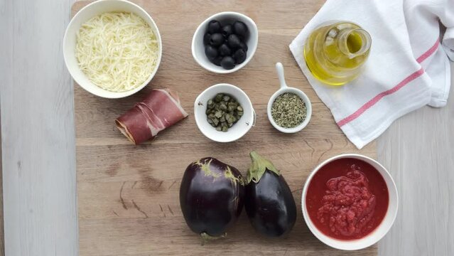 Mano de mujer coge un bol con r&uacute;cula de una tabla de madera con ingredientes de una receta de pizzas de berenjena.