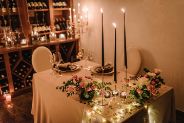 Dinner setting, date for two, Valentine's Day evening, burning candles. Candlelight date in a wine cellar. Glasses for wine, bouquet of flowers and decor on table. Romantic dinner at home at night.