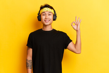 Young Asian man dressed in black t-shirt listening music via black wireless headphones, showing ok...