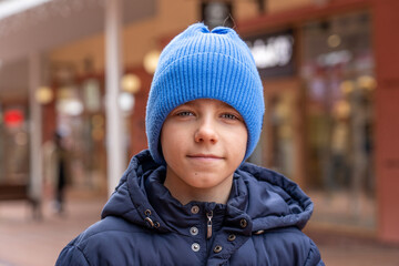 Portrait of confident teen boy in warm coat and blue hat looks directly into camera at city street.