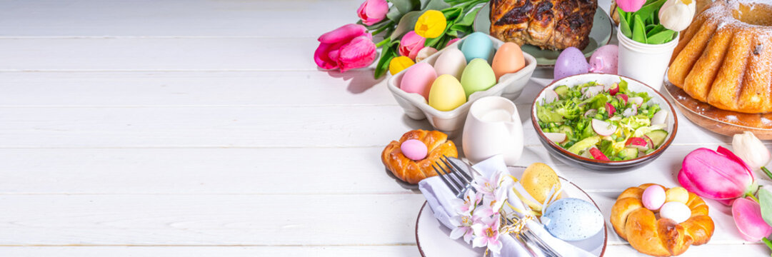 Festive served Easter dinner or brunch table, with Easter egg, tulip flower bouquet, traditional Easter foods - cake, cross buns, wreath bread, glazed ham and spring salad, white table background