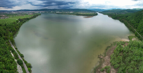 Naklejka premium Aerial drone panorama above a lake in Valcea County, Romania. Stormy weather, rain clouds are gathering above the surrounding forests. Several small villages can be seen on the lake's shores.