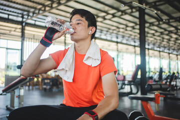 Fototapeta na wymiar Asian man is sitting on bench and drinking water to relax after workout in gym.