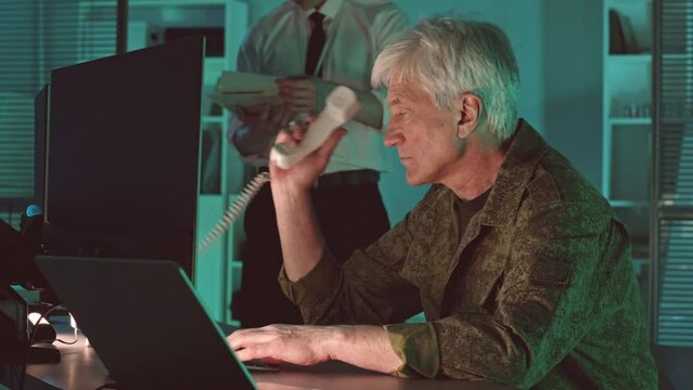 Senior Caucasian military surveillance commander in camouflage uniform making phone call while tracking target on computer screen working in dark control room