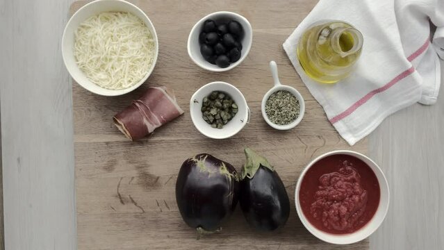 Ingredientes de receta salen de plano con mano de mujer. Aceite, aceitunas negras, queso rallado, jamón serrano, alcaparras, orégano, rúcula, berenjenas y tomate triturado.