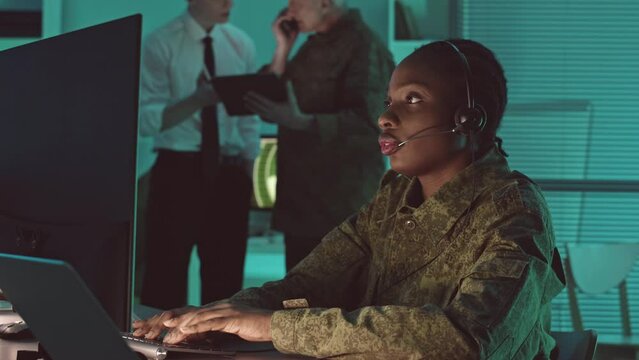 Young African American female military surveillance office in camouflage monitor monitoring cameras from satellite of target, controlling operation and making phone call, working in dark office hub